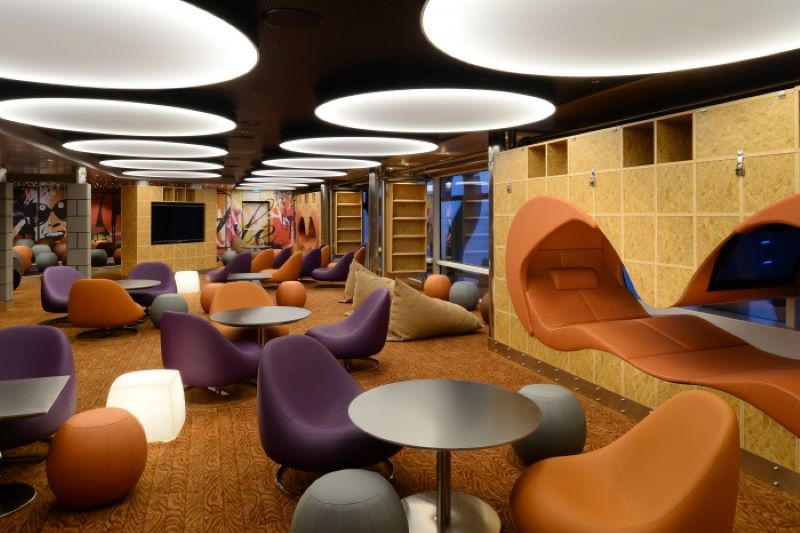 The Living Room on Royal Caribbean's Quantum of the Seas presents a trendy teen lounge with circular overhead lighting, funky purple and orange chairs, and abstract art, creating a cool and inviting space for young cruisers to hang out and relax.