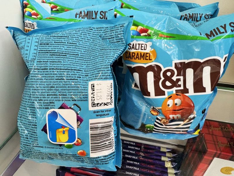 Bags of family-size Salted Caramel M&M's on display aboard a Cunard cruise ship, with the front and back of the packaging visible, including nutritional information and a colorful M&M character illustration.