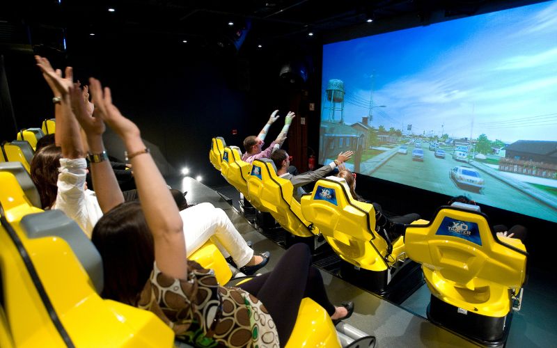 Excited passengers with raised hands enjoy a dynamic experience in the 4D Cinema on MSC Meraviglia, seated in yellow motion chairs that match the action on the large screen, depicting a lively street scene for an immersive entertainment activity.