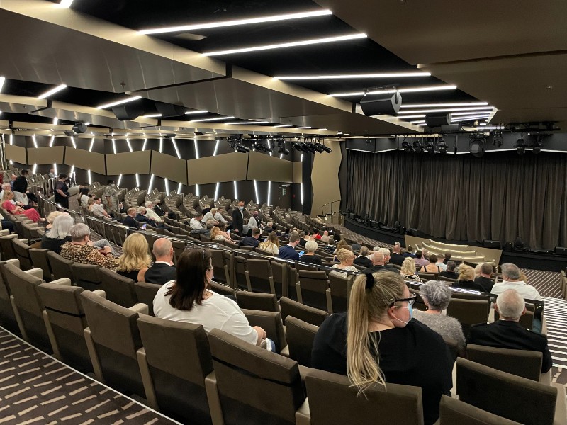 Passengers are seated in the contemporary theater on MSC Virtuosa, waiting for a performance to begin. The theater features modern lighting and a muted color palette, creating an inviting atmosphere for entertainment.