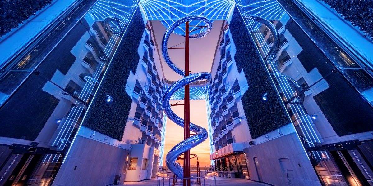 The 'Venom Drop' on MSC World Europa at twilight, an enthralling cruise ship waterslide that winds between LED-lit balconies, juxtaposing modern design with the thrill of an at-sea adventure, captured against a backdrop of a fading sunset.