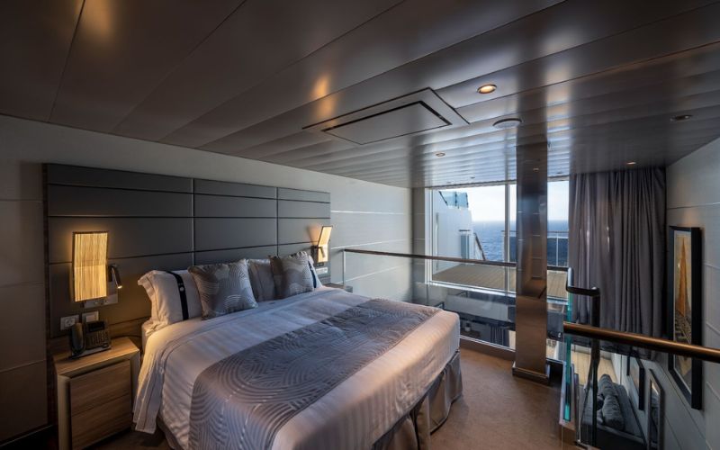 Inside the MSC Yacht Club Duplex Suite, showcasing a modern bedroom with a king-size bed, plush grey bedding, and floor-to-ceiling windows leading to a private balcony overlooking the ocean, offering luxury and comfort at sea.