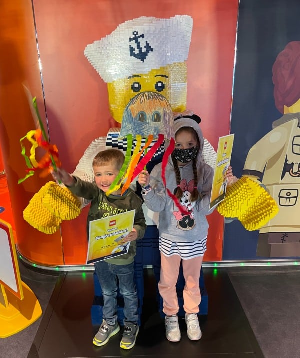 Two joyful children hold colorful paper crafts and a 'Captain' certificate in the MSC Cruises Kids Club, with a backdrop of a LEGO-style pirate captain, capturing the fun and creative spirit of the ship's youth activities.