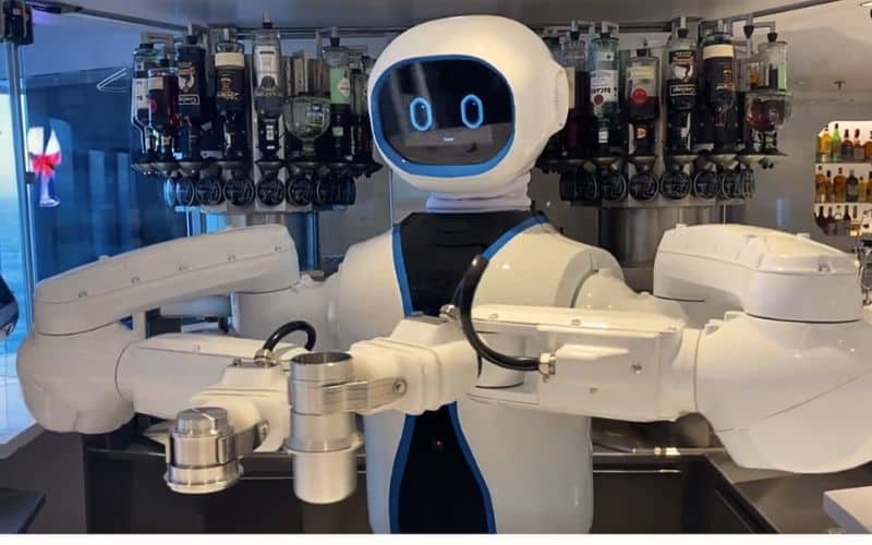 A robot bartender, featuring a friendly digital face and white mechanical arms, stands ready to mix drinks at a bar fully stocked with a variety of liquors and spirits on an MSC cruise ship, showcasing modern automation at sea.