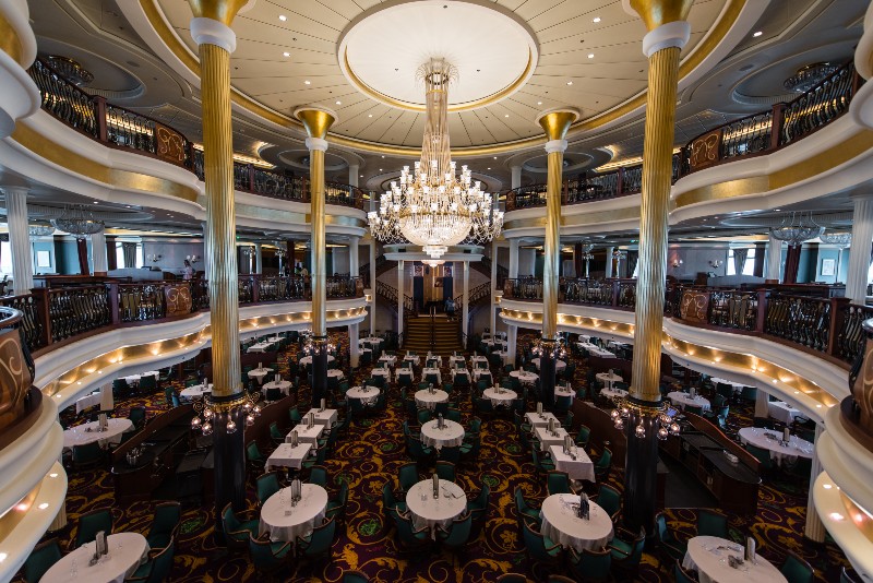 The grandeur of the Main Dining Room aboard Liberty of the Seas, highlighted by a striking chandelier, golden pillars, and elegant, circular tables set for a fine dining experience.