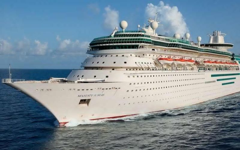 Royal Caribbean's 'Majesty of the Seas' cruise ship, easily identified by its name on the hull, cuts through the blue waters, topped with green-striped decks under a partly cloudy sky.