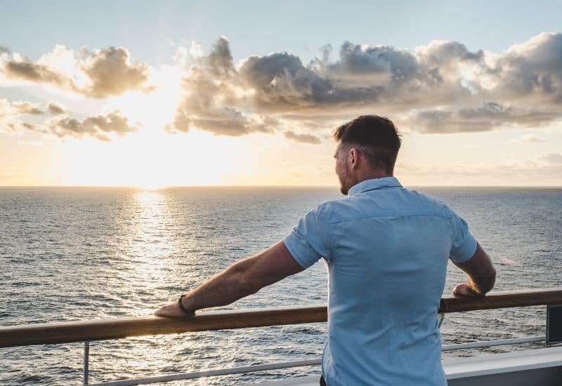 A man in a light blue shirt leaning on the deck railing of a cruise ship, looking out towards the horizon as the sun sets over the ocean.