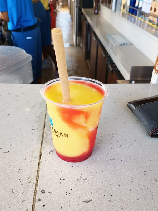A Mango Meltdown cocktail on a Norwegian Cruise Line ship, featuring a vibrant mix of yellow and red slush in a branded plastic cup with a straw. The drink is placed on a countertop at the bar, with bartenders in blue uniforms visible in the background.