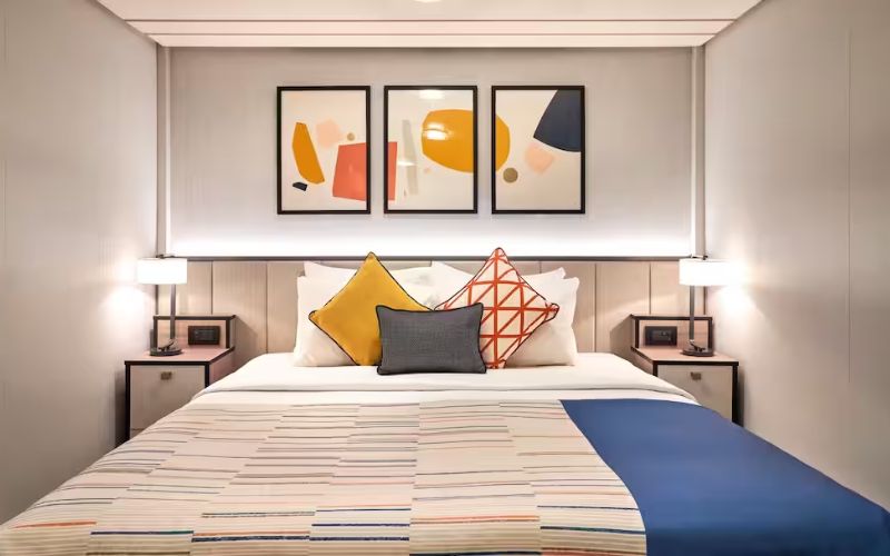 Modern cruise ship stateroom featuring a neatly made bed with striped bedding and vibrant geometric throw pillows, flanked by nightstands with lamps, and abstract art pieces above the bed headboard.