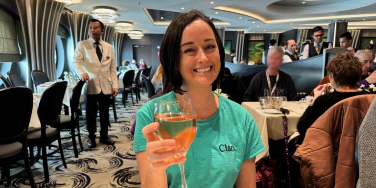 Cruise Mummy raises a glass of amber-colored beverage in the Main Dining Room on an MSC cruise ship, with elegant décor and attentive staff in the background, capturing a moment of joy and fine dining on a luxury voyage.