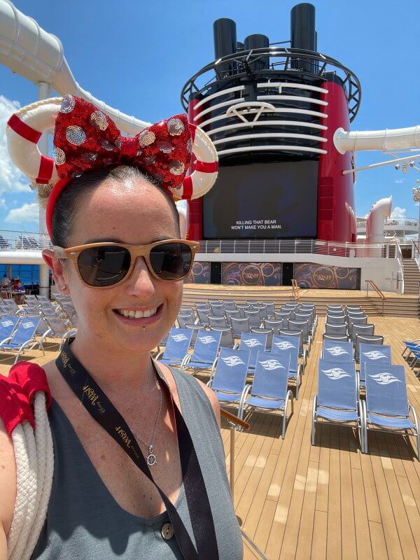 Cruise Mummy wearing sunglasses and a red polka dot Minnie Mouse bow headband smiles on the pool deck of Disney Cruise Line's Disney Wish. Behind her, lounge chairs are neatly arranged in front of a large screen on the ship's funnel, with clear blue skies overhead.