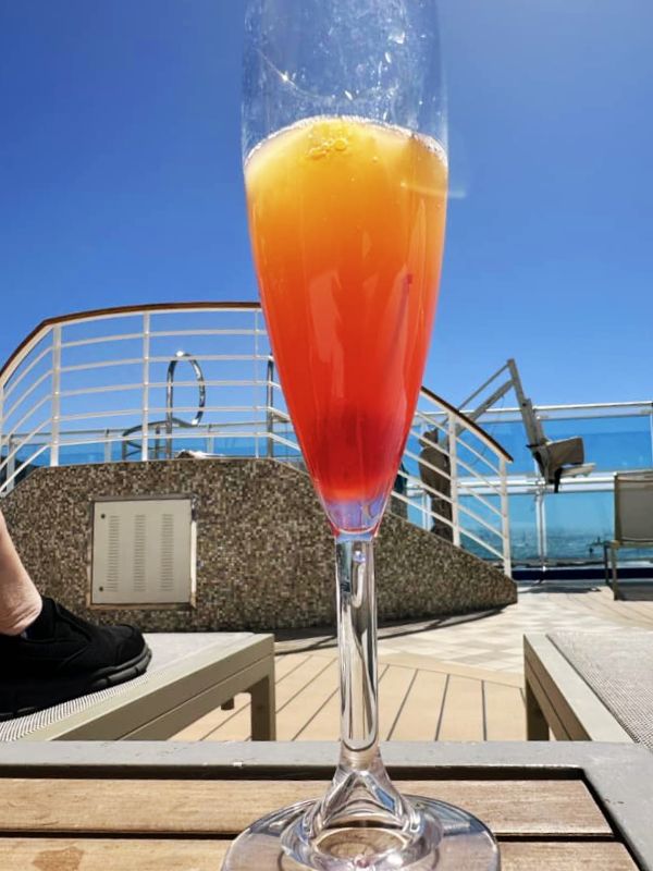 A Mermaid Mimosa on the Sky Princess cruise ship, served in a tall champagne flute with a gradient from red to orange. The drink is set on a deck table with a sunny blue sky and part of the ship's railing and deck area in the background, creating a bright and relaxing ambiance perfect for a day at sea.