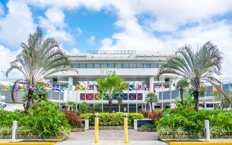 The image shows the exterior of Miami International Airport, featuring a modern building with a variety of international flags displayed prominently along the front. The scene is framed by lush green tropical plants and palm trees, with a bright blue sky and scattered clouds above, creating a welcoming atmosphere.