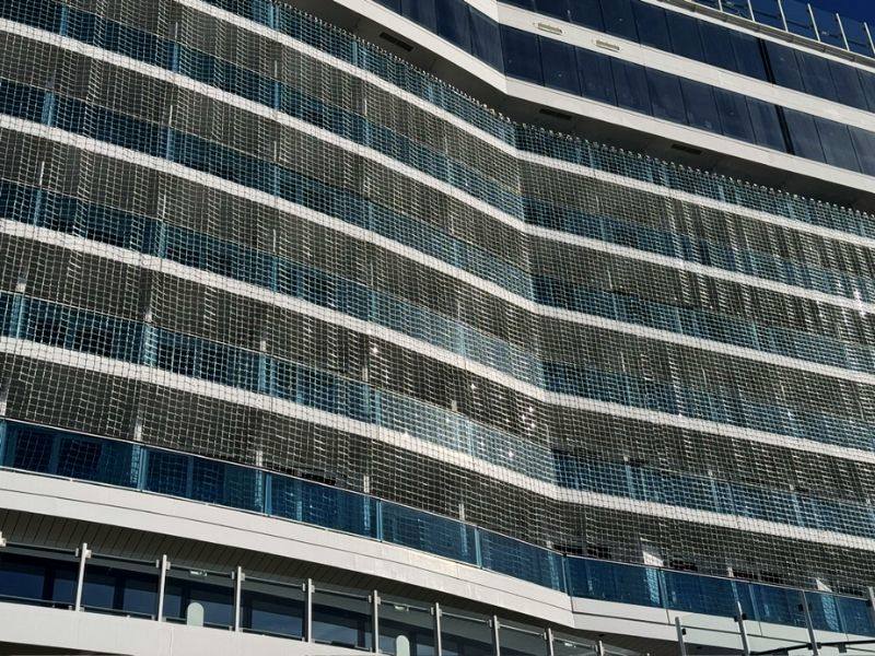 Close-up view of the midsection of the Costa Smeralda cruise ship, partially obscured by a large net, with a focus on the ship's blue-tinted balcony rooms and white exterior.