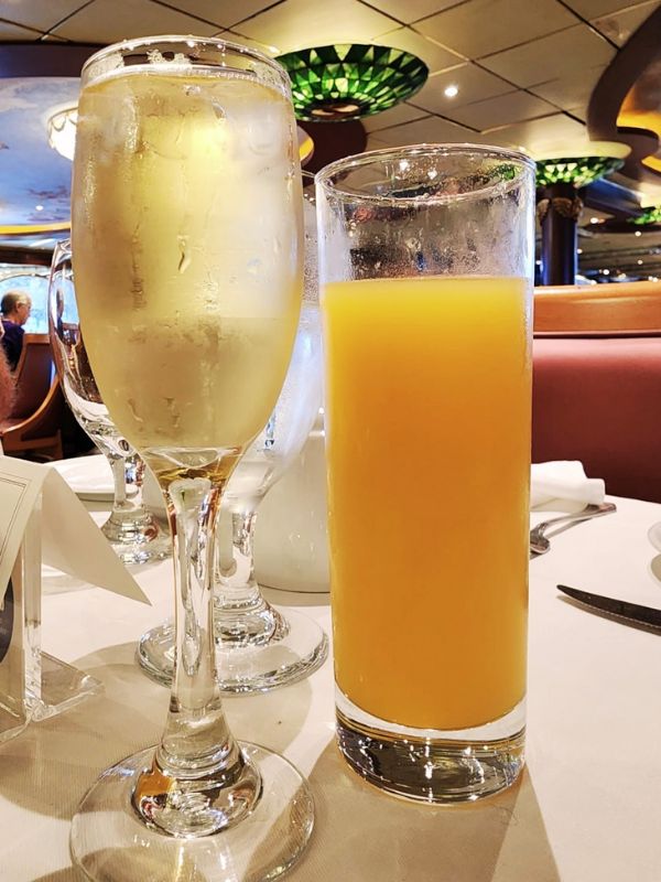 A mimosa served in a champagne flute alongside a tall glass of orange juice, set on a white tablecloth in a dining area on a cruise ship. The background features elegant lighting fixtures and seating, contributing to a sophisticated brunch setting.