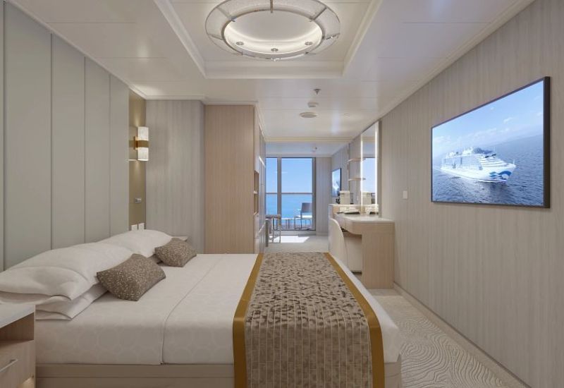 The image shows a Mini-Suite on Princess Cruises' Sun Princess. The suite features a spacious bed with luxurious linens, decorative pillows, and a gold-accented bed runner. A large flat-screen TV is mounted on the wall opposite the bed, displaying an image of a Princess cruise ship. The room extends to a private balcony, visible through sliding glass doors, offering outdoor seating and a view of the ocean. The decor is elegant with neutral tones and sophisticated lighting, creating a luxurious and comfortable ambiance. The suite also includes a desk area and ample space for relaxation.
