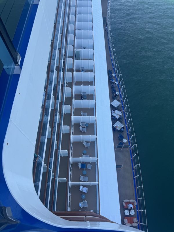 The image shows an aerial view of the Cabana Mini-Suites on Decks 9 and 10 of the Sun Princess. The balconies of the mini-suites are arranged in a row, with each featuring a small table and chairs, providing a private outdoor space for guests. The balconies are separated by white partitions for privacy. Below the balconies, there is a wider deck area with additional seating and tables, likely part of a public or shared outdoor space on the ship. The view highlights the ship's clean, modern design and the close proximity of the balconies to the water, offering guests a direct connection to the ocean.
