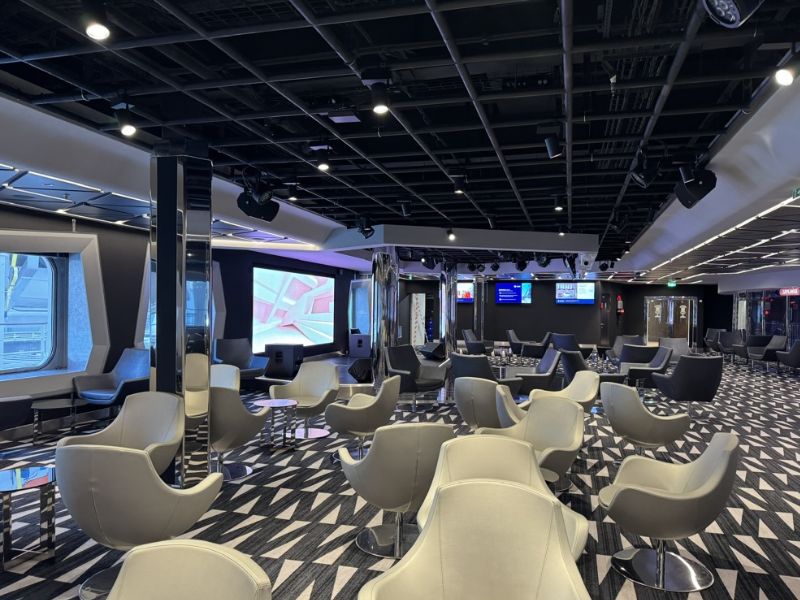 Modern and stylish interior of a cruise ship lounge with comfortable grey swivel chairs, geometric patterned carpet, and contemporary lighting. Large screens and windows suggest a space designed for entertainment and relaxation.