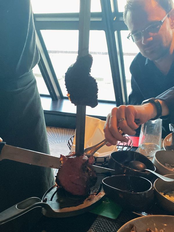 A person slicing a skewered piece of succulent meat at Moderno restaurant on NCL Getaway, capturing the essence of Brazilian steakhouse dining with a view of the ocean through the window.