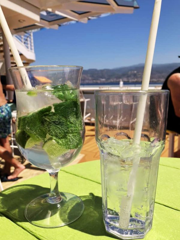 A mojito cocktail in a stemmed glass filled with mint leaves and lime wedges, placed next to an empty tall glass with a straw on a green table aboard a cruise ship. The background features a sunny deck area with passengers and scenic views of mountains and the ocean.