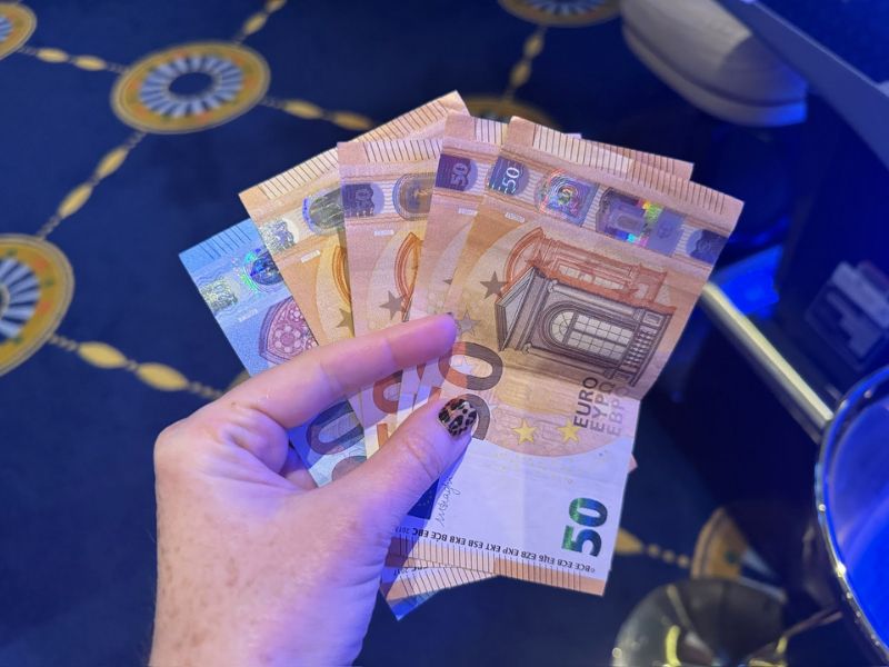 A hand holding several 50 Euro banknotes with blurred casino carpet in the background, indicating recent winnings from a casino on the Costa Smeralda cruise ship.