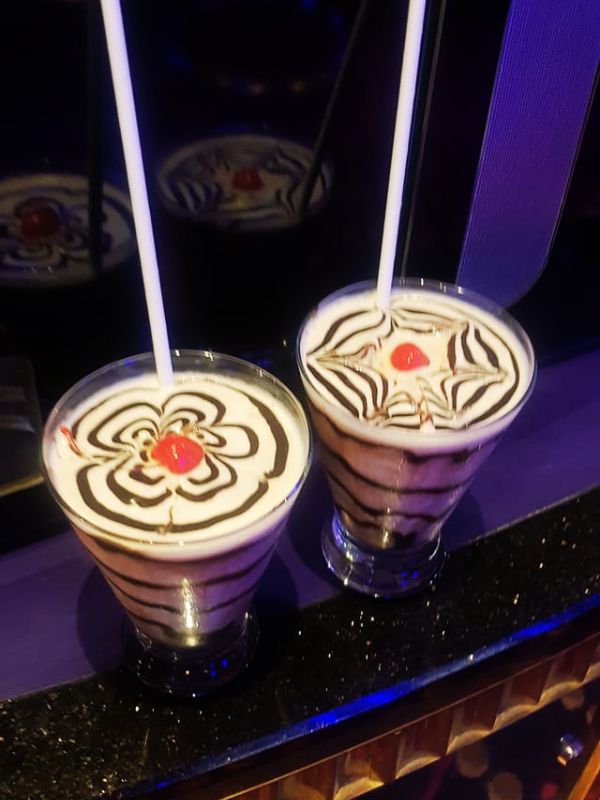 Two Mudslide cocktails on a cruise ship, topped with intricate chocolate syrup designs and a cherry in the center. The drinks are served in clear glasses with straws, and their reflection can be seen in a mirrored surface nearby, enhancing the visual appeal.