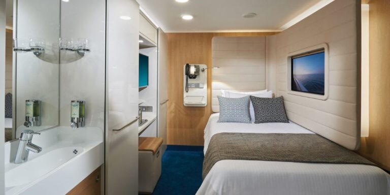 A modern Bliss Studio cabin on an NCL cruise ship, featuring a comfortable bed with a gray blanket and decorative pillows. The room includes a sleek bathroom area with a sink and toiletries, a wall-mounted TV, and a compact desk area, all designed with clean lines and neutral colors to maximize space and comfort.