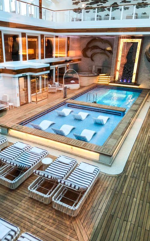 The courtyard of a Norwegian Cruise Line ship, showcasing an inviting pool with blue lights and partially submerged loungers, flanked by rows of striped cushioned deck chairs on a wooden deck, with multi-level balconied suites providing a view of the serene, upscale area.