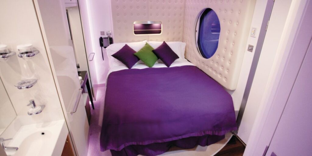 Interior of a Norwegian Cruise Line Epic studio cabin featuring a full-size bed with purple and green accent pillows, a round porthole window, and an integrated bathroom sink and shelving unit to the left.