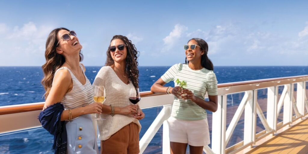 Three women sharing a joyful moment on a cruise ship's deck, with two holding glasses of wine and one with a cocktail. They are casually dressed, enjoying the open ocean view under a clear blue sky, reflecting a relaxed and luxurious cruise experience.