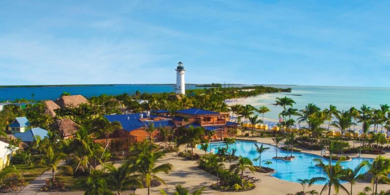 This image features an aerial view of Norwegian Cruise Line's Harvest Caye, showcasing its tropical resort-style setting. The scene includes a large pool surrounded by palm trees, colorful buildings, and a white lighthouse overlooking pristine beaches and turquoise waters. The vibrant and tranquil atmosphere highlights this private island destination's appeal to cruise visitors.