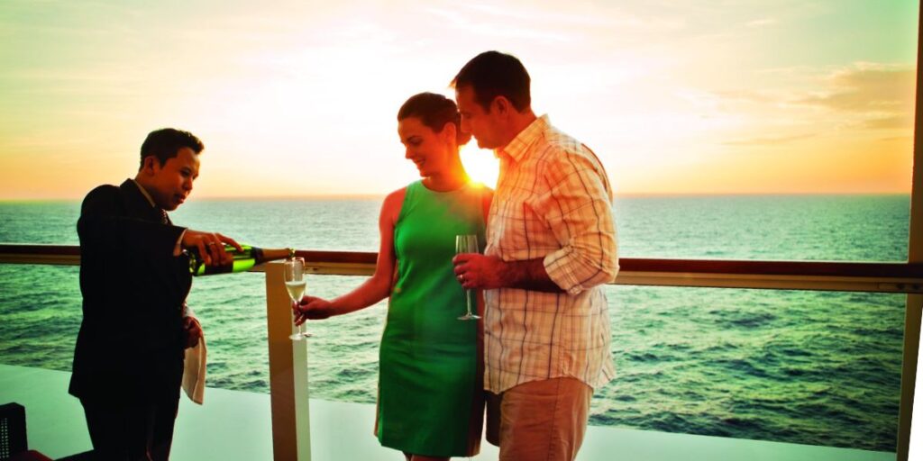 A waiter in a formal suit pours champagne into a glass for a couple enjoying a sunset on NCL cruise ship balcony, with a serene ocean horizon in the background.