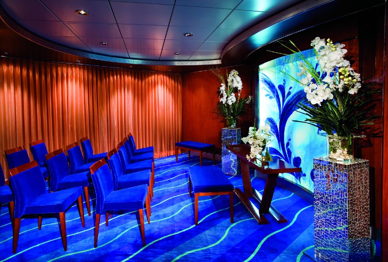 A small, elegant wedding chapel on NCL Pearl with blue velvet chairs arranged in rows facing a modern altar. The altar is flanked by two large floral arrangements and a blue-tinted decorative glass backdrop. The room is adorned with wood paneling and blue patterned carpet, creating a serene atmosphere.