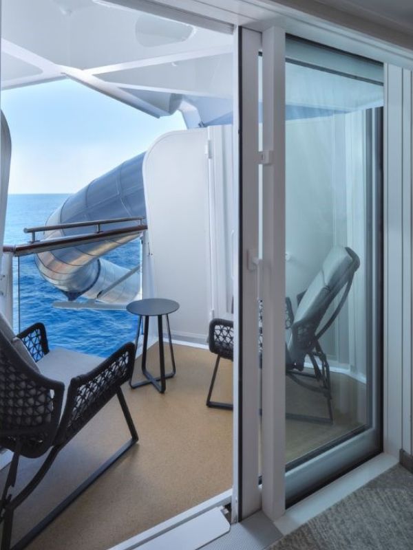 View from inside a Norwegian Prima stateroom with an ADA-accessible obstructed ocean view balcony, featuring comfortable outdoor furniture and a clear glass door, with a glimpse of a lifeboat outside.