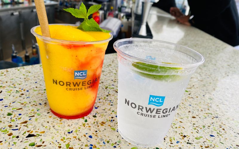 Two refreshing drinks on a cruise ship bar, with a vibrant yellow and red frozen cocktail garnished with mint in a cup labeled 'NORWEGIAN CRUISE LINE,' next to a clear drink with a lime wedge, showcasing the NCL's 'Free at Sea' indulgent beverage options.