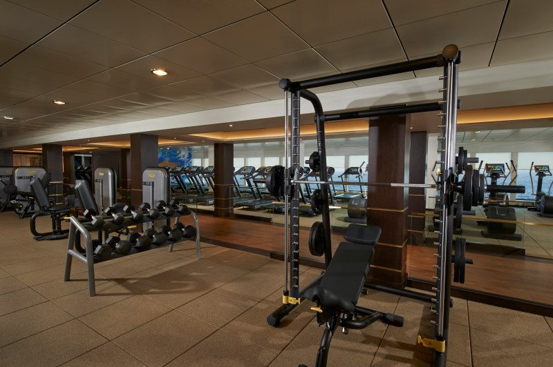 Well-equipped gym on a Norwegian Cruise Line ship, featuring a range of fitness equipment including treadmills, elliptical machines, free weights, and a Smith machine, with large windows offering ocean views.