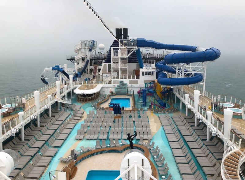 Overcast view of the Norwegian Encore's pool deck featuring multiple pools, lounging areas, water slides, and a climbing structure, with the ocean extending to the horizon.