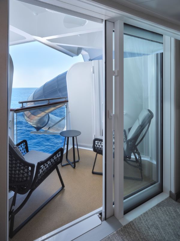 View from inside a Norwegian Prima stateroom with an ADA-accessible obstructed ocean view balcony, featuring comfortable outdoor furniture and a clear glass door, with a glimpse of a lifeboat outside.