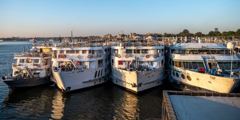 Several Nile cruise ships moored side by side at a dock in the golden hour of the early evening. The ships are multi-decked with rows of windows and life rings, bustling with activity against a backdrop of the distant shoreline under a clear sky. This scene captures the vibrant atmosphere of river cruising in Egypt.