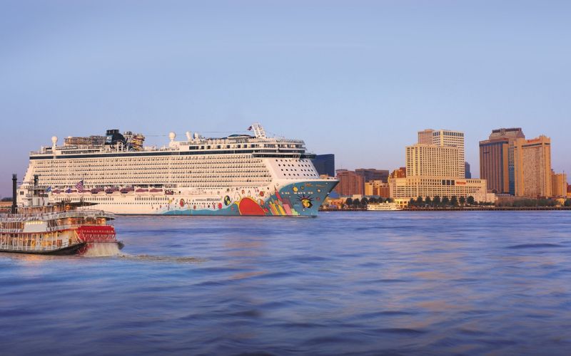 This image shows the Norwegian Breakaway cruise ship sailing on a calm body of water near New Orleans. The ship is large and decorated with vibrant artwork on its hull. In the background, you can see the New Orleans skyline, including several tall buildings, under a clear, light blue sky. A smaller riverboat is also visible on the left side of the image, adding to the scenic view of the city and the river.