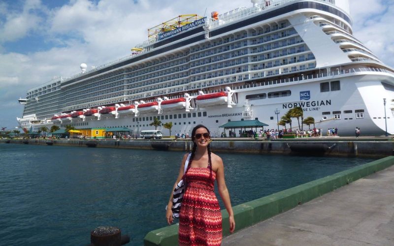 Cruise Mummy in a red and white summer dress stands smiling at the dockside with the Norwegian Breakaway cruise ship moored in the background, under a bright blue sky with some clouds.