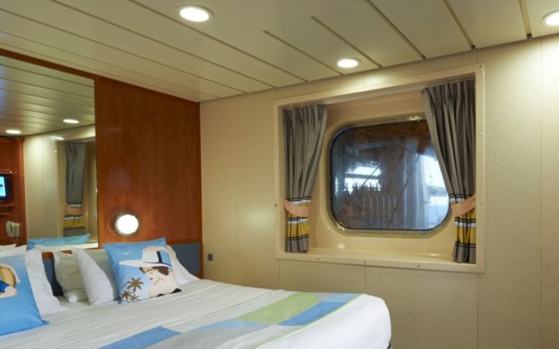 A cozy stateroom on the Norwegian Dawn with an obstructed ocean view, featuring a large bed with colorful striped linens, maritime-themed pillows, and a porthole window partially obscured by lifeboat equipment.