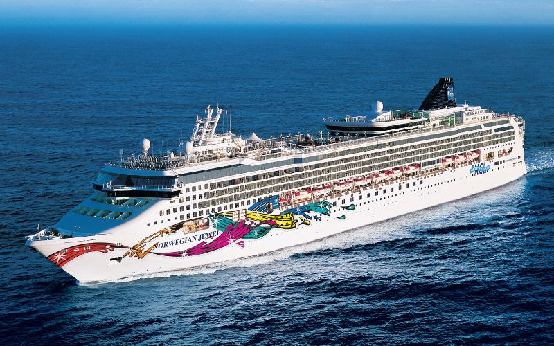 The Norwegian Jewel cruise ship cuts through the deep blue sea, its hull adorned with colorful, vibrant artwork. The ship, captured in bright sunlight, showcases the leisurely luxury of ocean travel against the vast expanse of the ocean.