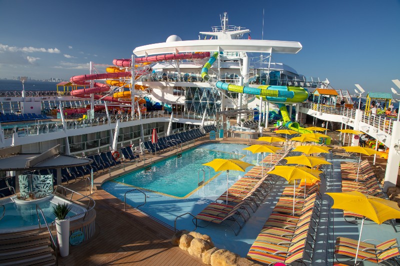 Sunny pool deck on the Oasis of the Seas with vibrant yellow umbrellas, colorful waterslides, and loungers, set against a clear blue sky