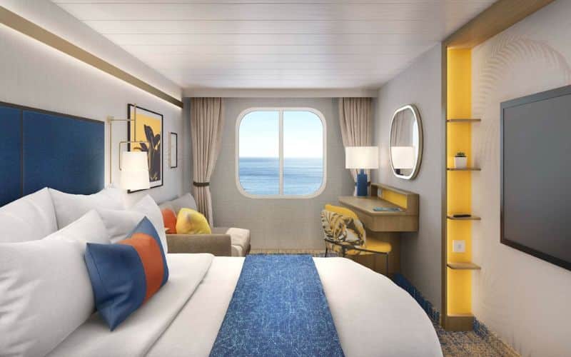 Elegantly appointed ocean view stateroom on Royal Caribbean's 'Icon of the Seas,' featuring a large bed with pristine white linens and vibrant blue and orange accent pillows. A large porthole window offers a serene view of the ocean, complemented by stylish furnishings including a beige sofa, a flat-screen TV, and a small work area with modern amenities.