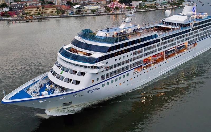 The Oceania Regatta cruise ship sails gracefully near a city waterfront during dusk, its lights beginning to twinkle against the settling evening sky.