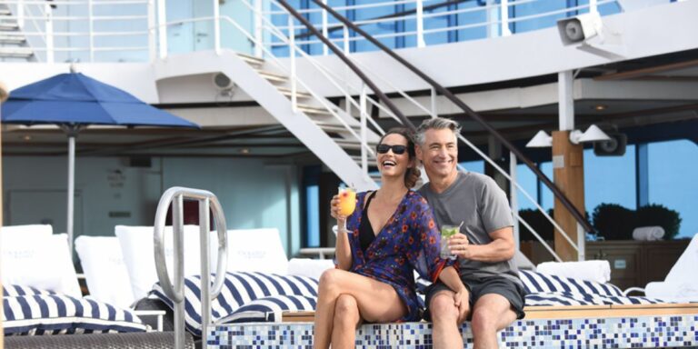 A joyful couple lounging on deck chairs by the poolside of a cruise ship, sharing a laugh over refreshing drinks, with the sunny ambiance reflecting a relaxing and happy vacation moment.