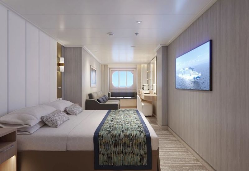 The image shows Oceanview stateroom on Princess Cruises' Sun Princess. The room features a large bed with a patterned bed runner and multiple pillows, positioned to face a window with an ocean view. The stateroom includes a wall-mounted flat-screen TV displaying an image of a cruise ship, a sofa, and a desk area with a mirror and coffee machine. The decor is modern with soft, neutral tones, emphasizing comfort and a serene ambiance, perfect for relaxing while enjoying the ocean scenery.