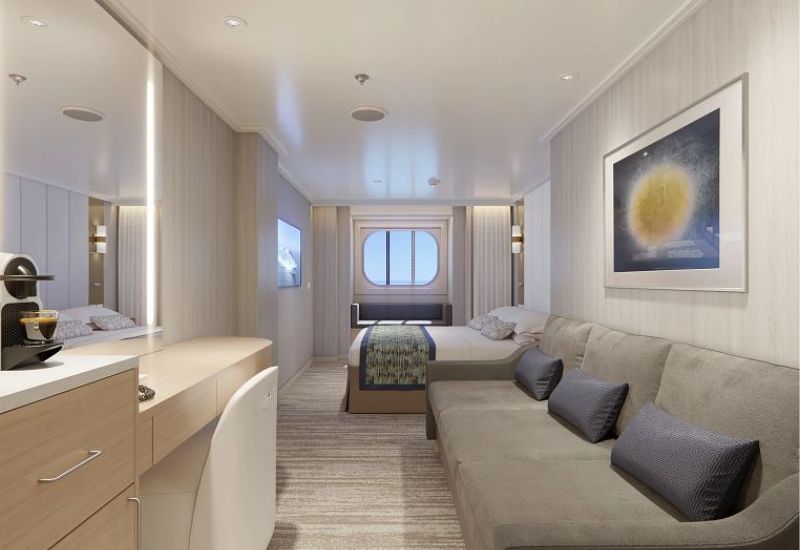 The image depicts an Oceanview stateroom on Princess Cruises' Sun Princess. The room features a large window that offers a view of the ocean, with a bed positioned to take advantage of the scenery. The decor is modern and light, with a neutral color palette. The room includes a sofa with accent pillows, a desk with a mirror, and a coffee machine. A framed artwork adds a touch of elegance to the space, and the overall design emphasizes comfort and luxury.