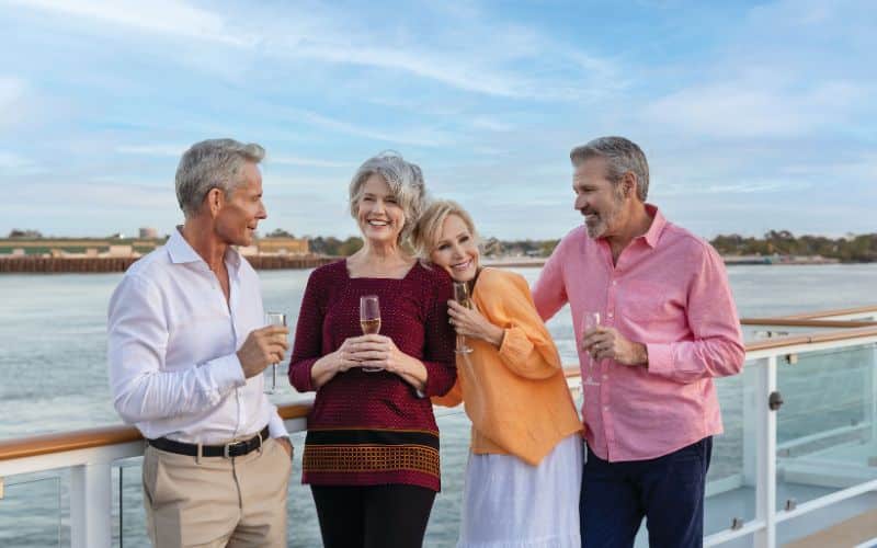Two mature couples enjoying a moment together on a cruise ship deck, each holding a glass of champagne. They are smiling and engaged in a cheerful conversation against a backdrop of a tranquil waterway and a distant shoreline. The setting is casual yet elegant, ideal for conveying leisure and companionship among friends.